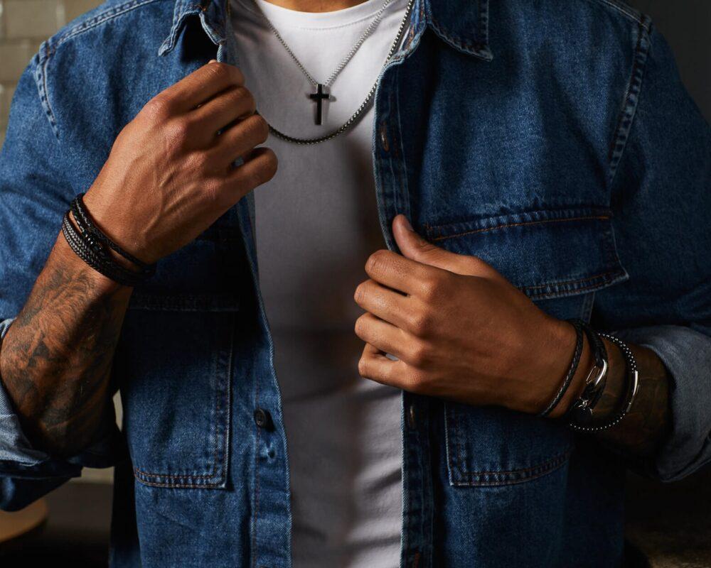 Man wearing denim shirt, with bracelets on both wrists and multiple necklaces.